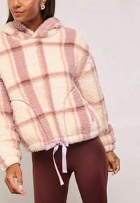 Rózsaszín és krémszínű kockás flísz kapucnis pulóver, húzózsinóros aljjal, lila szalaggal, és oldalsó zsebekkel. Puha, bolyhos textúra.