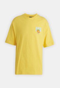 Camiseta de algodón amarilla con un corte relajado. Presenta un gráfico de un limón de dibujos animados y la palabra "NUEVO" en azul en la zona del bolsillo frontal.