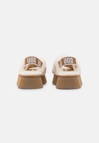 Zapatillas de casa beige suaves con una suela de plataforma marrón claro, que cuentan con una tira ancha con el logo de UGG en blanco en la parte posterior.