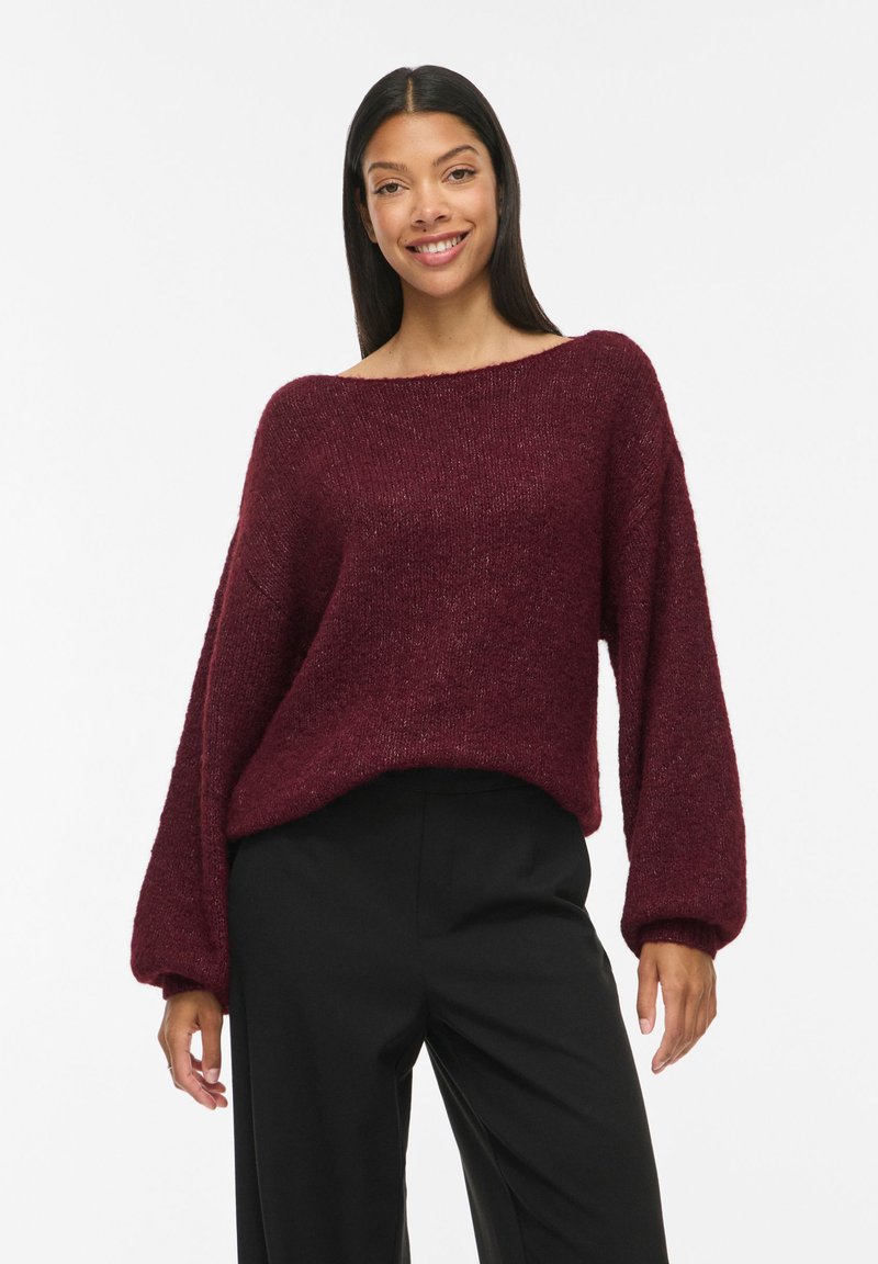 Femme aux longs cheveux foncés portant un pull ample bordeaux et un pantalon noir, souriante devant un fond blanc uni.