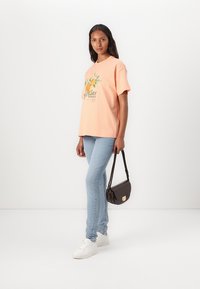 Camiseta de color naranja claro con estampado floral, de corte relajado, combinada con jeans azul claro. Un bolso negro tipo bandolera y zapatillas blancas completan el look.