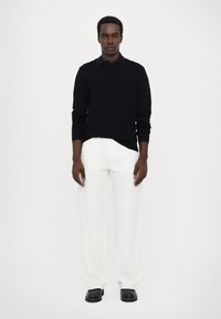 Pull en tricot noir avec un motif en losange, associé à un pantalon large blanc et des chaussures noires, se tenant devant un fond blanc uni.