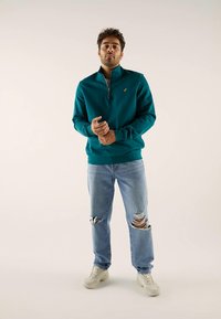 Hombre vistiendo un sudadera de media cremallera en color verde azulado, jeans claros desgastados y zapatillas blancas, de pie contra un fondo claro liso.