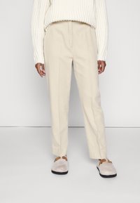 Beige, rechte broek van zachte stof, met een plooi aan de voorkant, gecombineerd met lichtgrijze instappers met bruine accenten.
