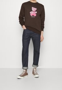 Felpa marrone scuro con una grafica di un orsetto rosa, abbinata a jeans di denim scuro e scarpe alte marroni con suole bianche.