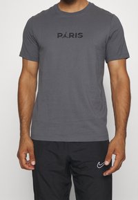 Grå t-shirt för män med kort ärm, med svart text "PARIS" och en logotyp på framsidan. Smidig bomullsstruktur, avslappnad passform.