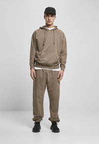 Felpa color khaki con tasca frontale e cordini, abbinata a pantaloni della tuta coordinati. Un berretto nero e sneakers scure completano l'outfit.