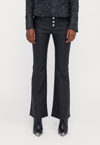 Zwarte uitlopende broek van een gladde, glanzende stof met een hoge taille en drie metalen knopen. Gecombineerd met puntige zwarte hoge hakken.