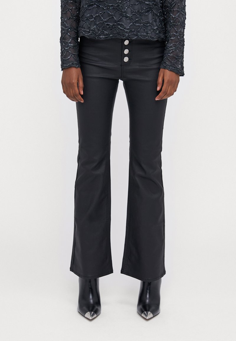 Zwarte uitlopende broek van een gladde, glanzende stof met een hoge taille en drie metalen knopen. Gecombineerd met puntige zwarte hoge hakken.