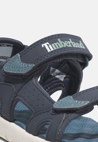 Tmavě modré sandály s nastavovacími pásky, texturovanou stélkou a kontrastními tyrkysovými akcenty. Logo Timberland je zobrazeno na pásku.