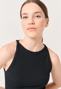 Schwarzes, geripptes Tanktop mit hohem Kragen und ärmellosem Design. Der Stoff wirkt glatt, körpernah, mit minimalen Nähten und ohne sichtbare Hardware.