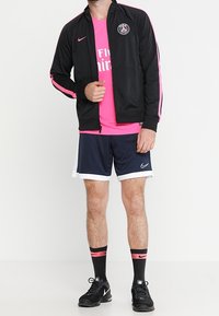 Schwarze Sportjacke mit pinken Akzenten, pinkes Shirt, navy Shorts, schwarze Sneaker und gemusterte Socken. Sportbekleidung, die für Komfort und Leistung ausgelegt ist.