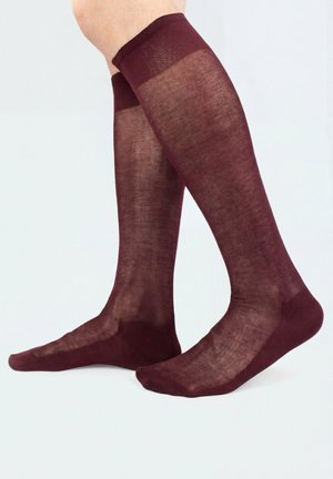 Benen met doorschijnende bordeaux kniekousen met verstevigde tenen en manchetten, staand op een lichte achtergrond.