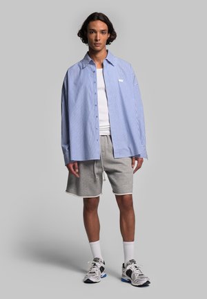 Blå og hvidstribet skjorte med knapper, grå shorts, hvide sportsstrømper og sneakers. Har et logo på brystet og en afslappet pasform.