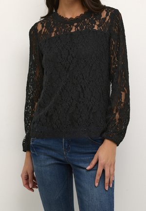Femme portant un top en dentelle noire à manches longues avec motif floral, associé à un jean en denim bleu, debout devant un fond uni.