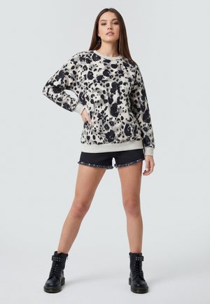 Jonge vrouw staand met de hand op de heup, draagt een zwart-witte trui met vlekken, zwarte short met franjes en zwarte combat boots op een lichte achtergrond.