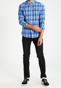 Persona che aggiusta la manica indossando una camicia a quadri blu con bottoni, pantaloni neri e scarpe sportive bianche e nere, in piedi su uno sfondo bianco.