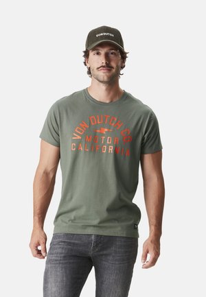 T-shirt olive à manches courtes avec un imprimé graphique rouge et orange "Von Dutch Co. Motor California" ; associé à un pantalon en jean foncé et une casquette assortie.