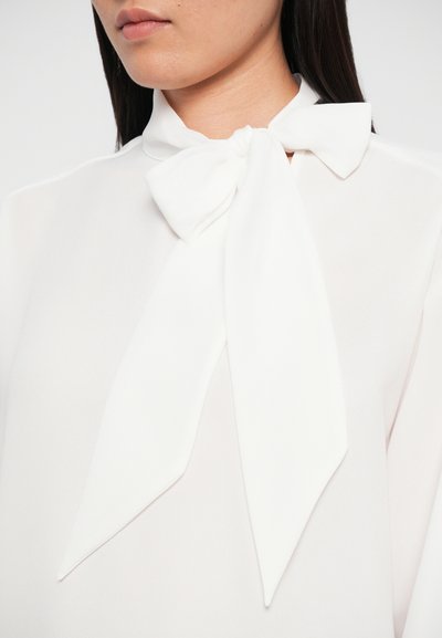 White blouse with a tie collar, featuring soft, smooth texture, long sleeves, and a loose fit. The tie is elegantly draped and knotted.