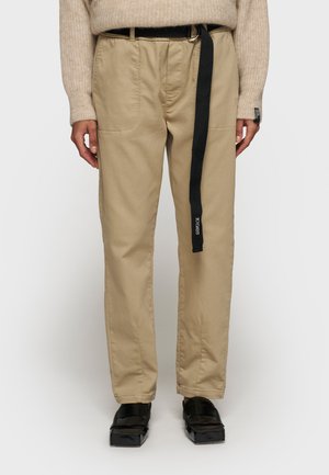 Khaki chinos met een ontspannen pasvorm, voorzien van voorzakken en een zwarte stoffen riem, gecombineerd met zwarte instapschoenen. Gladde katoenstructuur.