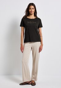 T-shirt en coton noir à col rond et texte imprimé, associé à un pantalon large beige et des chaussures slip-on en mesh noires.
