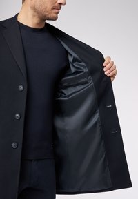 Cappotto nero su misura con una llining interna liscia, caratterizzato da grandi bottoni e una tasca interna, esposto aperto per rivelarne la struttura.