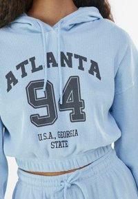 Ljusblå, texturerad hoodie med dragsko, med "ATLANTA 94" och "U.S.A. GEORGIA STATE" tryckt i svart. Croppad design.
