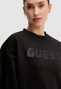 Felpa nera con logo "GUESS" in paillettes e texture sulla parte anteriore. Ha una scollatura tonda e maniche ampie per una vestibilità comoda.