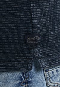 Cămașă din material tricotat în stil waffle de culoare bleumarin, având o etichetă mică albastră cu text alb. Cămașa are un hem curbat și este asortată cu blugi bleumarin deschis.