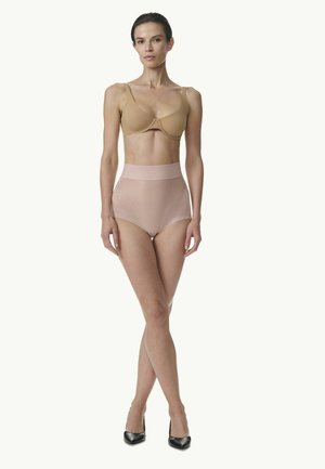 Frau steht und trägt einen beigen BH, hoch taillierte Nude-Formwäsche und schwarze High Heels vor einfarbigem Hintergrund.