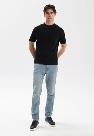Czarny bawełniany t-shirt z okrągłym dekoltem, w połączeniu z jasnoniebieskimi dżinsami. Model nosi czarne trampki z białymi podeszwami.