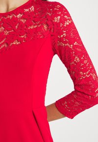 Robe rouge avec dentelle florale sur le haut de la poitrine et manches trois-quarts, avec un tissu uni sur le torse et une taille ajustée.