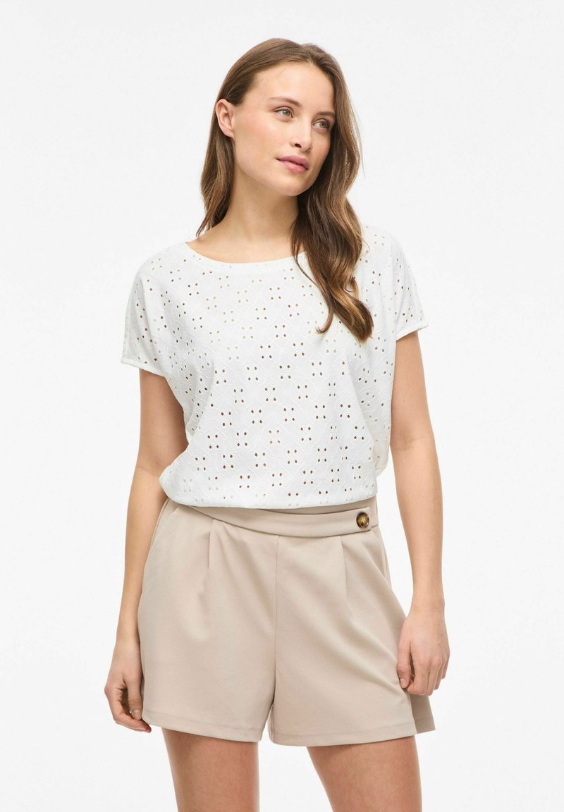 Femme aux longs cheveux bruns portant un haut blanc à manches courtes en œillet et un short beige taille haute avec détail boutons.