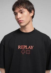 T-shirt nera oversize in cotone, con una stampa grafica rossa della parola "REPLAY" con accenti. Maniche corte e scollatura rotonda.