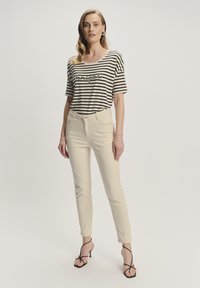 Pasiasta czarno-biała koszulka z krótkimi rękawami i grafiką, zestawiona z jasnobeżowymi dopasowanymi spodniami oraz czarnymi sandałami na obcasie.