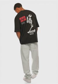 Sort oversized t-shirt med et hvidt skeletgrafik og rød tekst på ryggen; parret med lyse, løstsiddende bukser.
