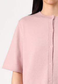 Cardigan corto rosa con maniche corte realizzato in morbido tessuto, caratterizzato da un colletto rotondo e chiusura frontale con cinque bottoni. Finitura texturizzata.