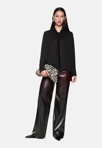 Schwarze Bluse mit langen Ärmeln und Kragen, kombiniert mit hochglänzenden burgunderroten weit geschnittenen Hosen. Hält eine Clutch mit Leopardenmuster.