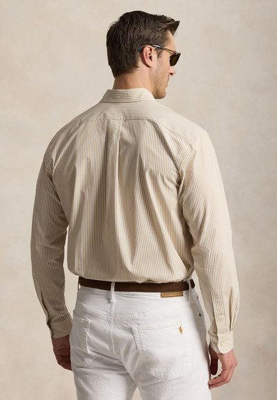 Man draagt een beige gestreept overhemd met lange mouwen, ingestopt in een witte broek met een bruine riem, met zijn rug naar de camera tegen een effen achtergrond.