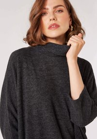 Př oversized tmavě šedý pletený svetr s vysokým límcem, texturovaným materiálem a tříčtvrtečními rukávy. Má volný střih a jemný pocit.