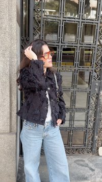 Moteris tamsiais plaukais, nešiojanti oranžinius akinius, su tekstūruotu juodu švarku ir šviesiai mėlynomis džinsomis, stovinti šalia puošnių geležinių ir stiklo durų.