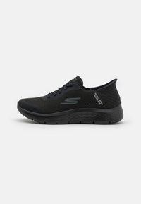 Zapatilla deportiva negra con parte superior de malla transpirable, acentos texturizados y suela acolchada. Presenta una punta redondeada y diseño con cordones.