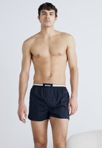 Shorts boxer noirs avec une ceinture contrastante arborant le nom de la marque "BOSS". Le tissu semble lisse avec une fermeture à bouton sur le devant.