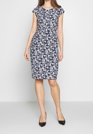 Frau trägt ein knielanges, marineblaues Kleid mit weißem Blumenmuster und kurzen Ärmeln, kombiniert mit schwarzen spitzen Pumps.