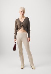 Maglione corto marrone con texture e nodo frontale, abbinato a pantaloni a coste color crema e scarpe beige a punta. Tiene una borsa color bordeaux.