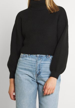 Woman wearing a black ribbed turtleneck sweater with balloon sleeves and light blue high-waisted jeans, standing against a plain background.