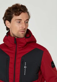 Röd och svart vadderad jacka med hög krage, fickor med dragkedja och en logotyp på bröstet. Har strukturerad tyg och kontrasterande färger.