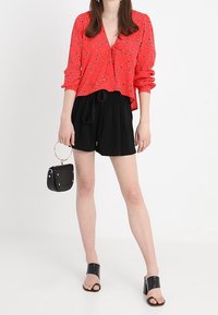 Czerwona bluzka w kwiaty z długimi rękawami, czarne szorty o wysokim stanie, czarne sandały na słupku oraz mała czarna torebka z złotymi akcentami.