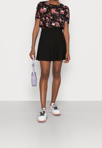 Blusa nera con fiori, gonna nera svasata e sneaker multicolore. Tiene una piccola borsa lavanda con una tracolla.