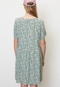 Mladá žena otočená zády, oblečená v pastelových květovaných šatech a růžové baseballové čepici, s dlouhými rovnými vlasy, na čistém bílém pozadí.